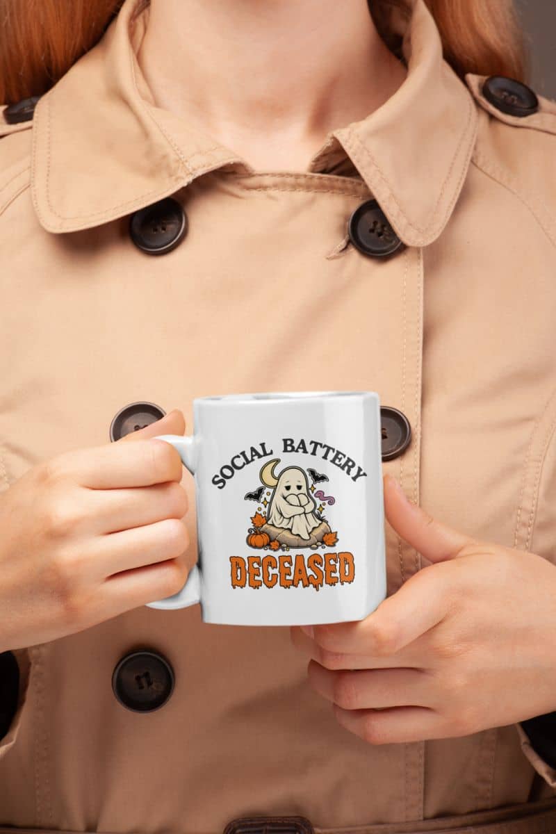 A person in a beige trench coat holding a spooky coffee cup with a 'Social Battery: Deceased' graphic in black and orange.