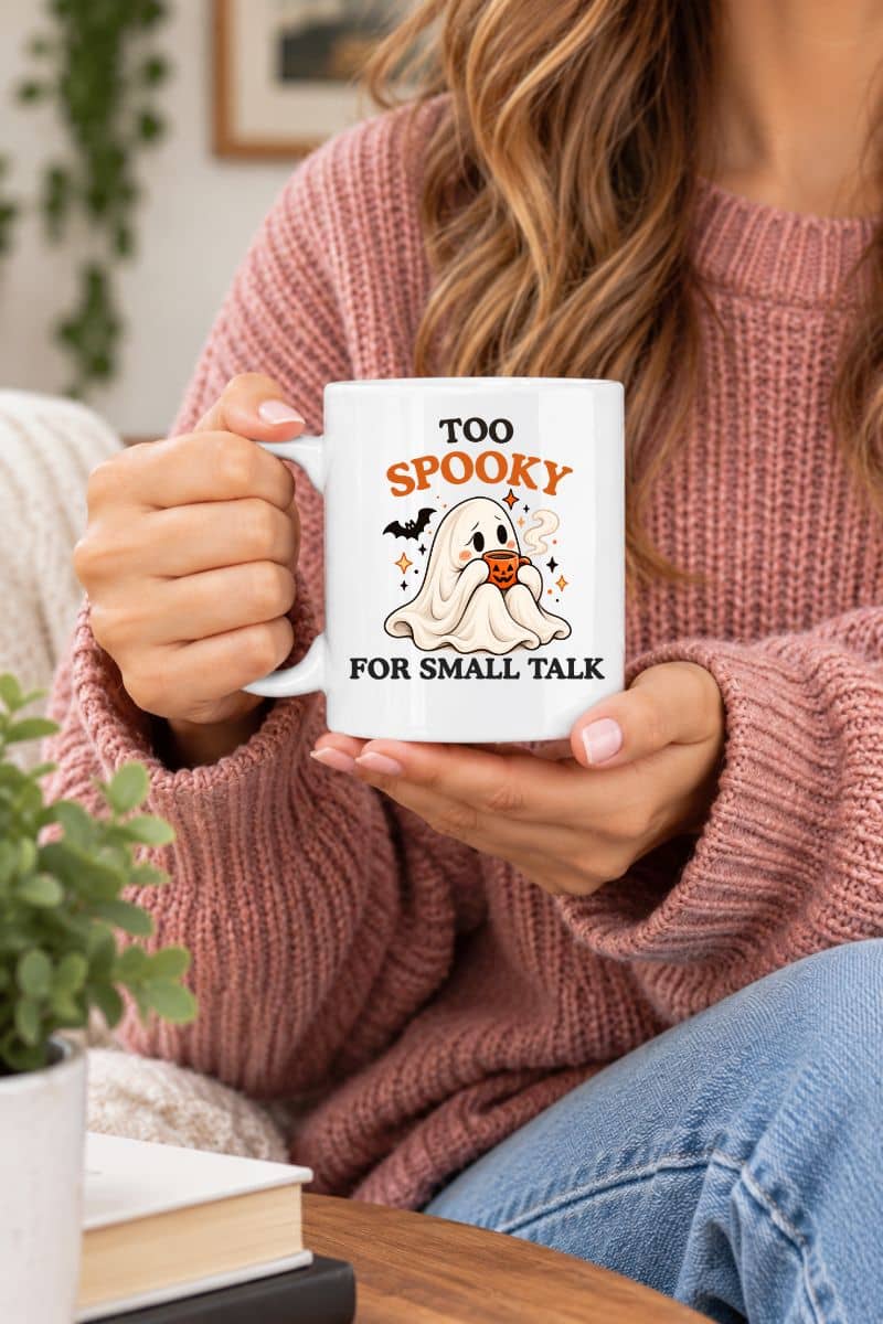 A woman in a pink knit sweater holds a spooky coffee mug featuring a coffee-loving ghost and the funny Halloween quote “Too Spooky for Small Talk.”