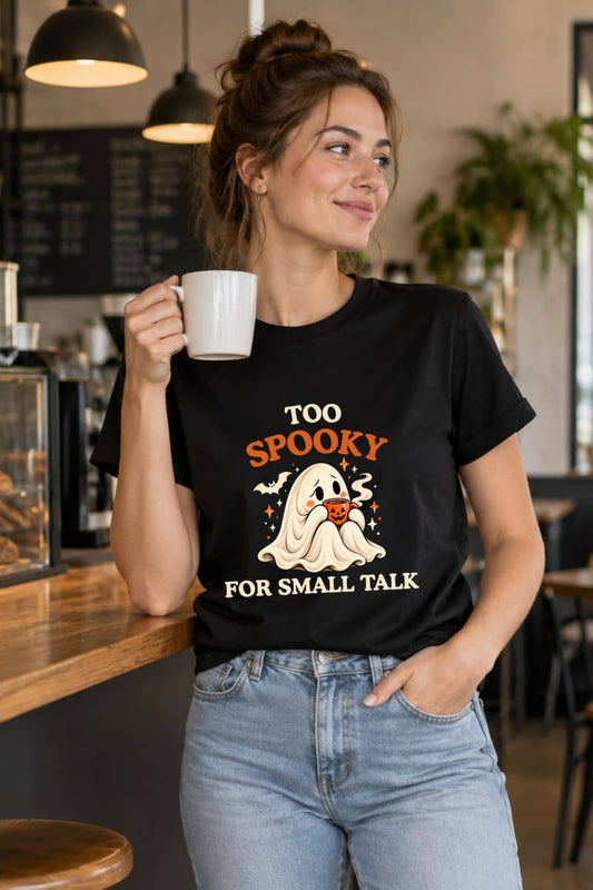 Woman wearing a black spooky season shirt featuring a shy ghost holding a pumpkin coffee mug, standing in a cozy café.