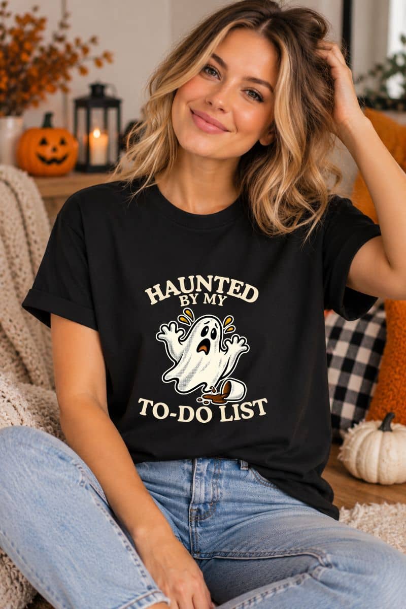 spooky shirts with a woman wearing a  black t-shirt featuring a “Haunted by My To-Do List” funny ghost, sitting on a sofa