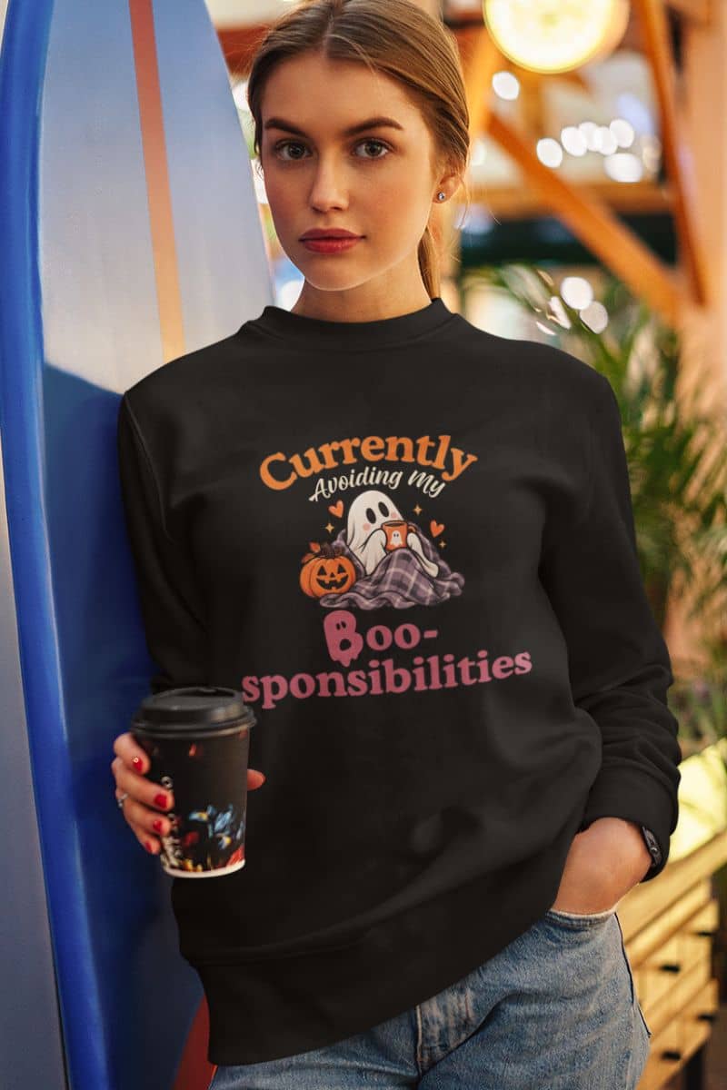 A woman wearing a spooky sweatshirt with a Halloween graphic that reads 'Currently Avoiding My Boo-sponsibilities' in orange and pink text, holding a coffee cup