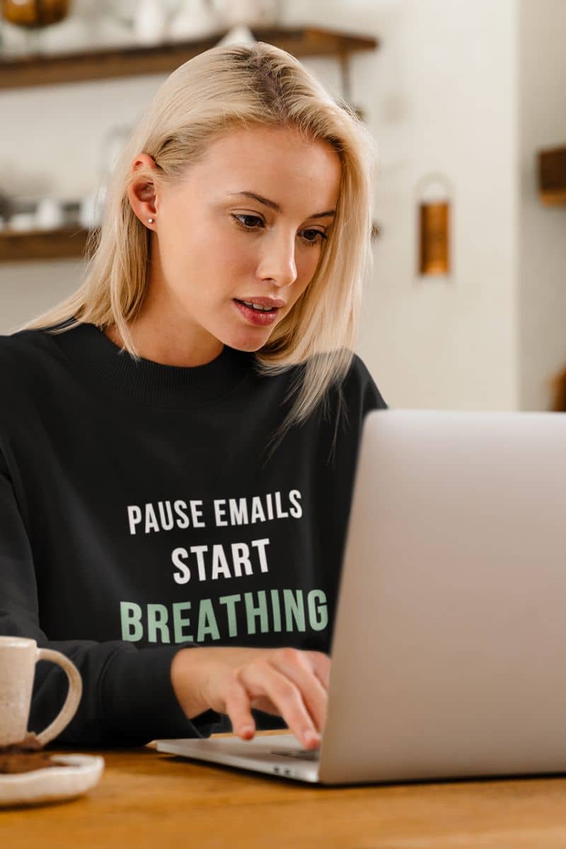stress relief pullover with a woman wearing a black sweatshirt with text 'Pause Emails, Start Breathing', using a laptop in a kitchen.