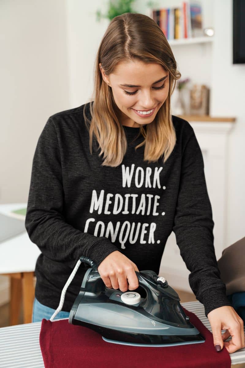 stress relief sweatshirt with a woman ironing a shirt with motivational text 'Work. Meditate. Conquer.' on her black sweatshirt.