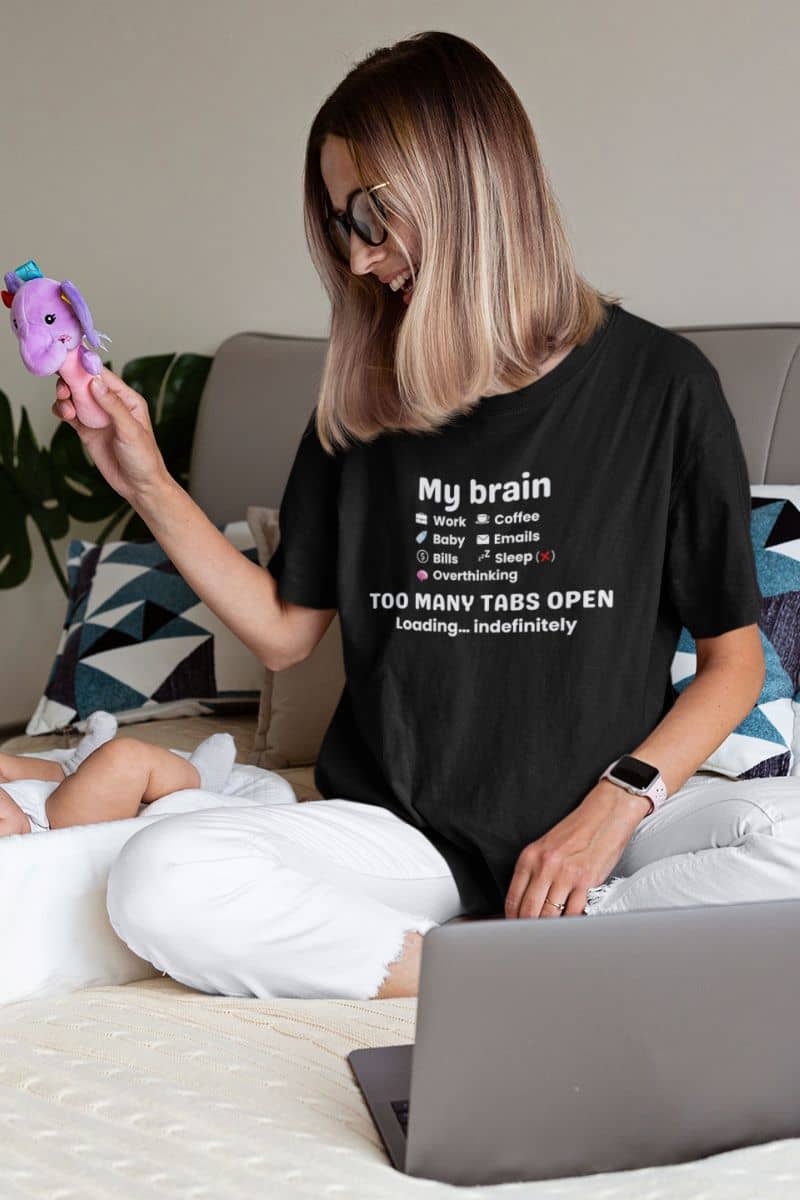 A stressed but funny shirt worn by a woman sitting on the bed, playing with her baby while working, featuring the text “My Brain Has Too Many Tabs Open.”