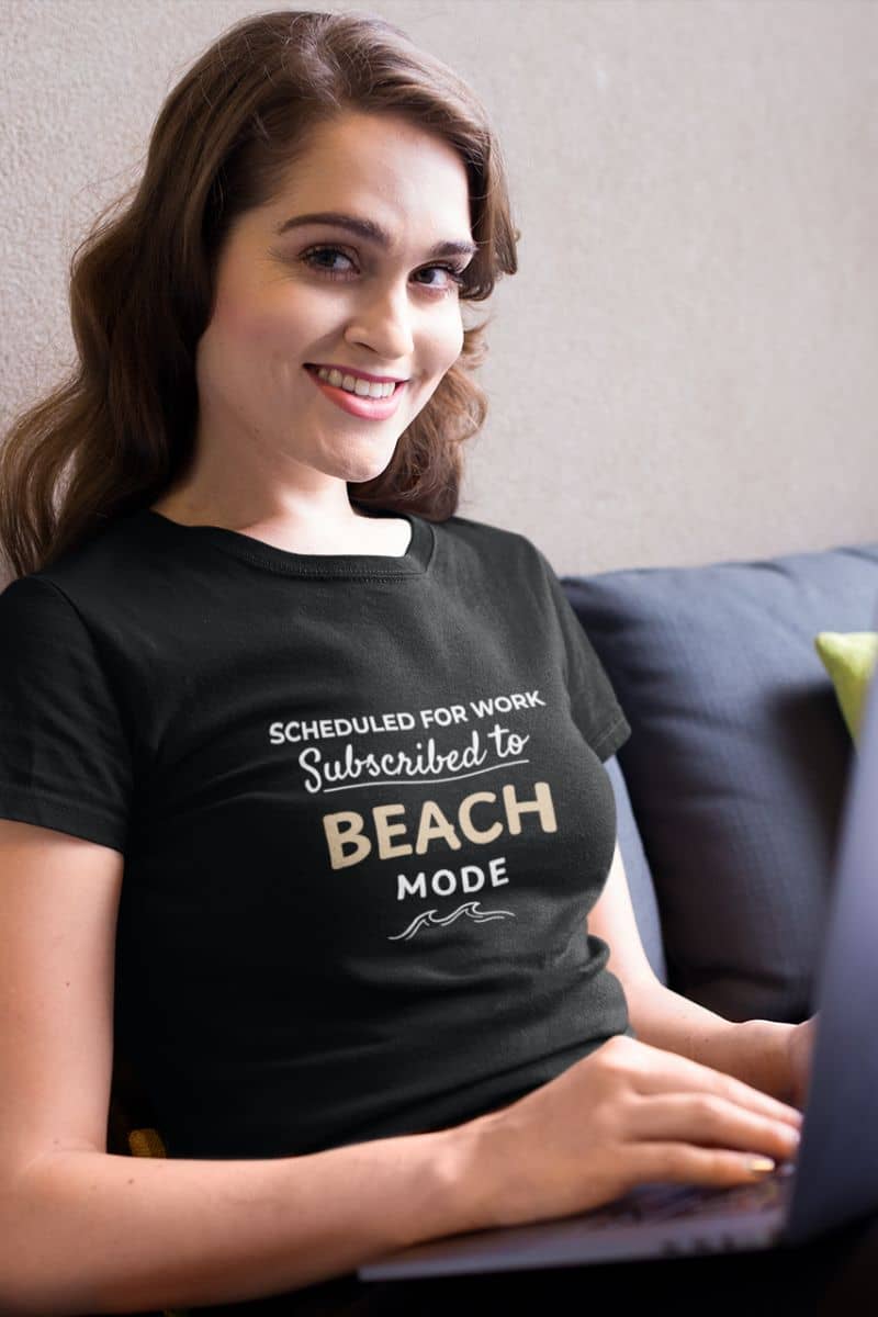 summer beach shirt with a woman wearing a black t-shirt with text 'Scheduled for Work. Subscribed to Beach', sitting on a couch with a laptop.