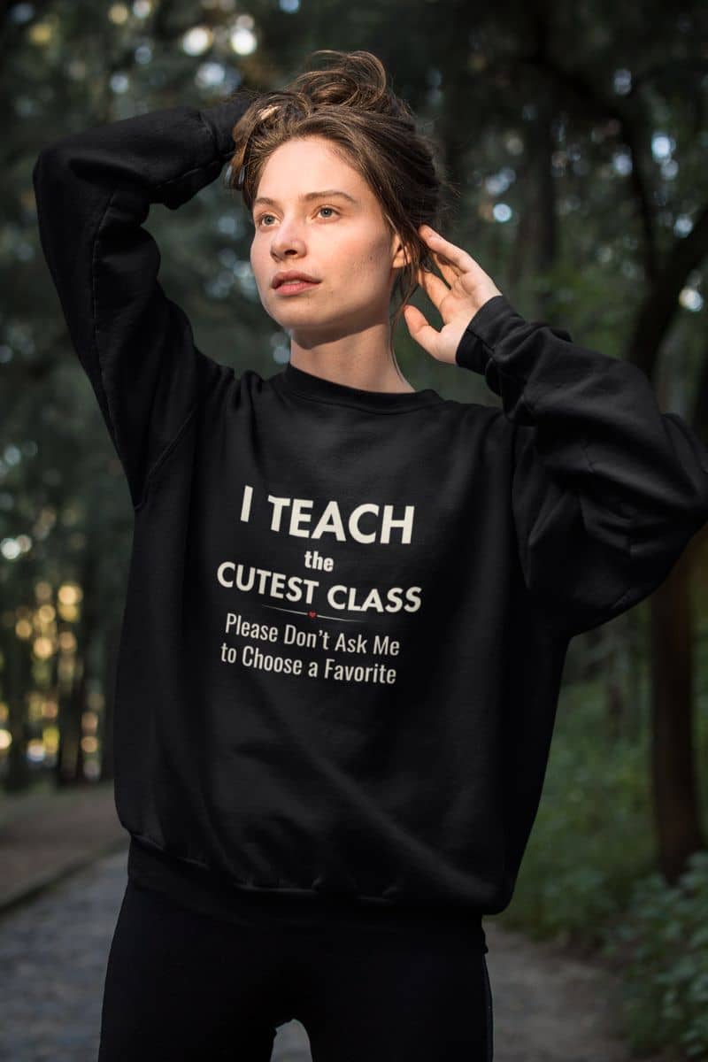 sweaters for teachers with a woman wearing a black crewneck sweatshirt featuring the text that reads 'I Teach the Cutest Class. Please Don’t Ask Me to Choose a Favorite', standing outdoors in a wooded setting.