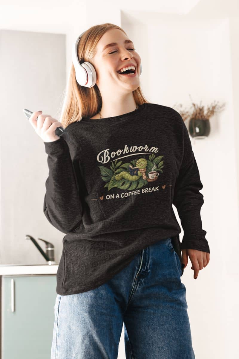 sweatshirt for readers with a woman wearing a dark charcoal heather graphic sweatshirt with a 'Bookworm on a Coffee Break' design, blue jeans, and white headphones, smiling in a bright kitchen setting.
