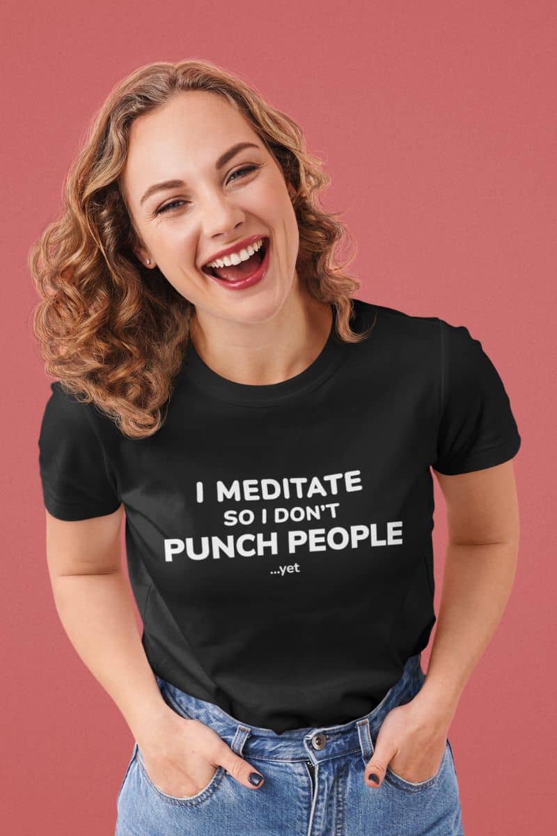 t shirts for introverts with a woman wearing a black t-shirt with humorous text 'I Meditate So I Don’t Punch People...yet' on a pink background