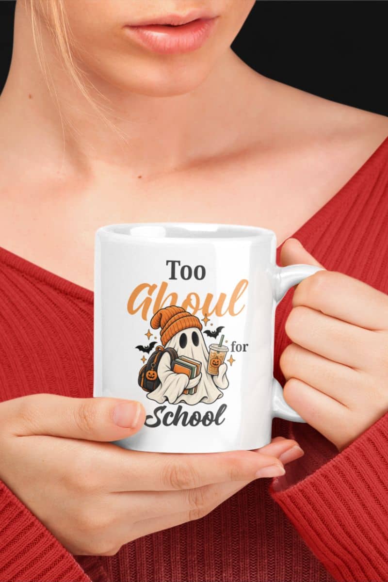 A person in a red ribbed sweater, holding a teacher Halloween mug with a Halloween-themed graphic that reads 'Too Ghoul for School' featuring a ghost holding books and coffee