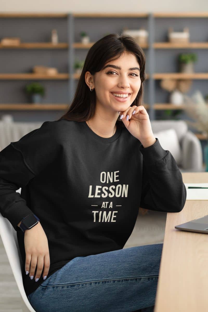 teacher appreciation gift with a woman wearing a black crewneck sweatshirt with the white printed text 'ONE LESSON AT A TIME' sits at a wooden desk in a modern office setting.