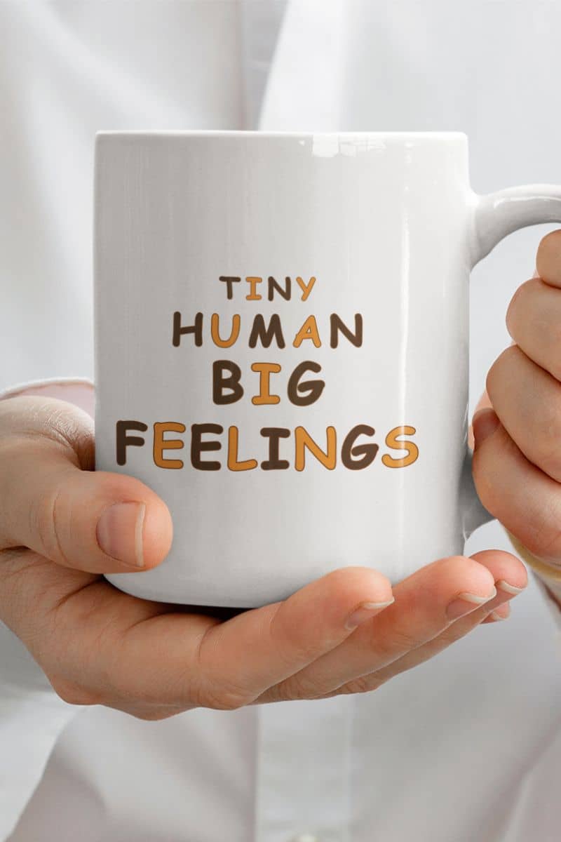 teacher appreciation mug with a white ceramic mug with the phrase 'TINY HUMAN BIG FEELINGS' printed on it, held in two hands against a white shirt background.