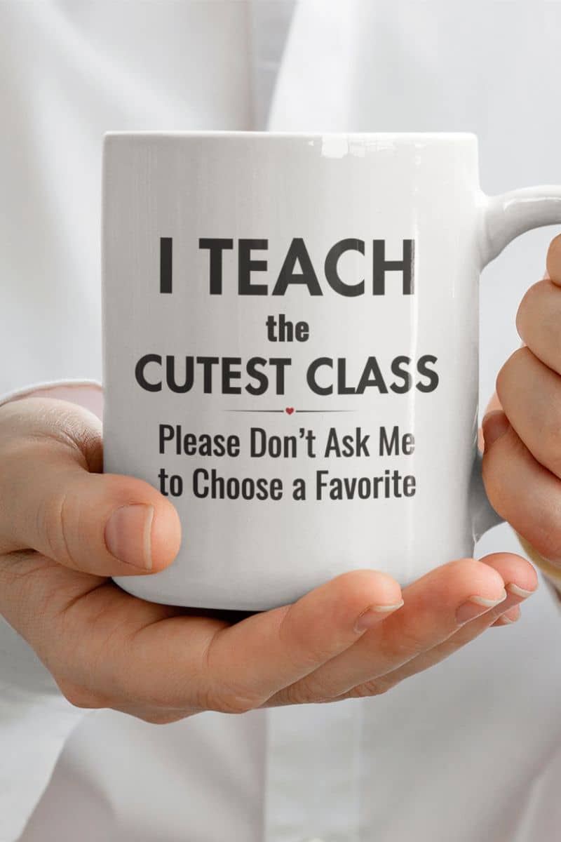 teacher appreciation mug held by a person, featuring the text 'I Teach the Cutest Class. Please Don’t Ask Me to Choose a Favorite', against a white shirt background.