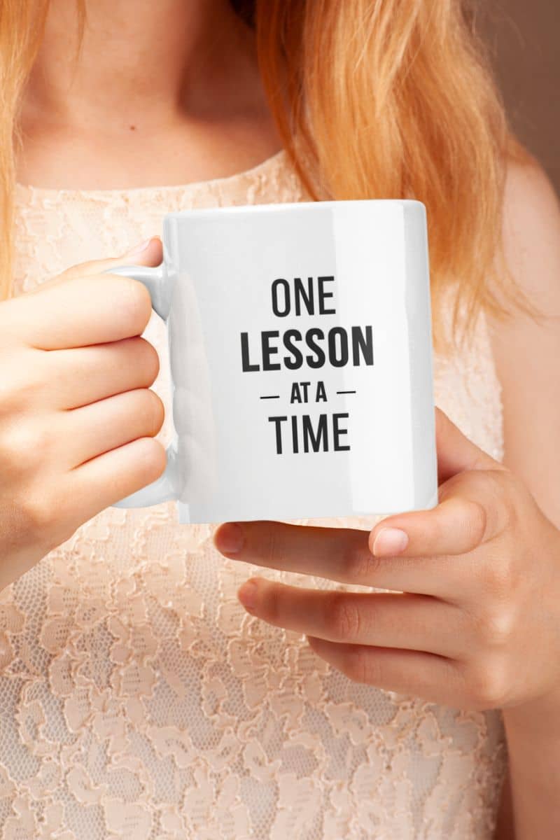 teacher appreciation mug with a person holding a white ceramic mug with the text 'ONE LESSON AT A TIME' printed in black. The person is wearing a light beige lace top, and the background is a soft, warm neutral tone.