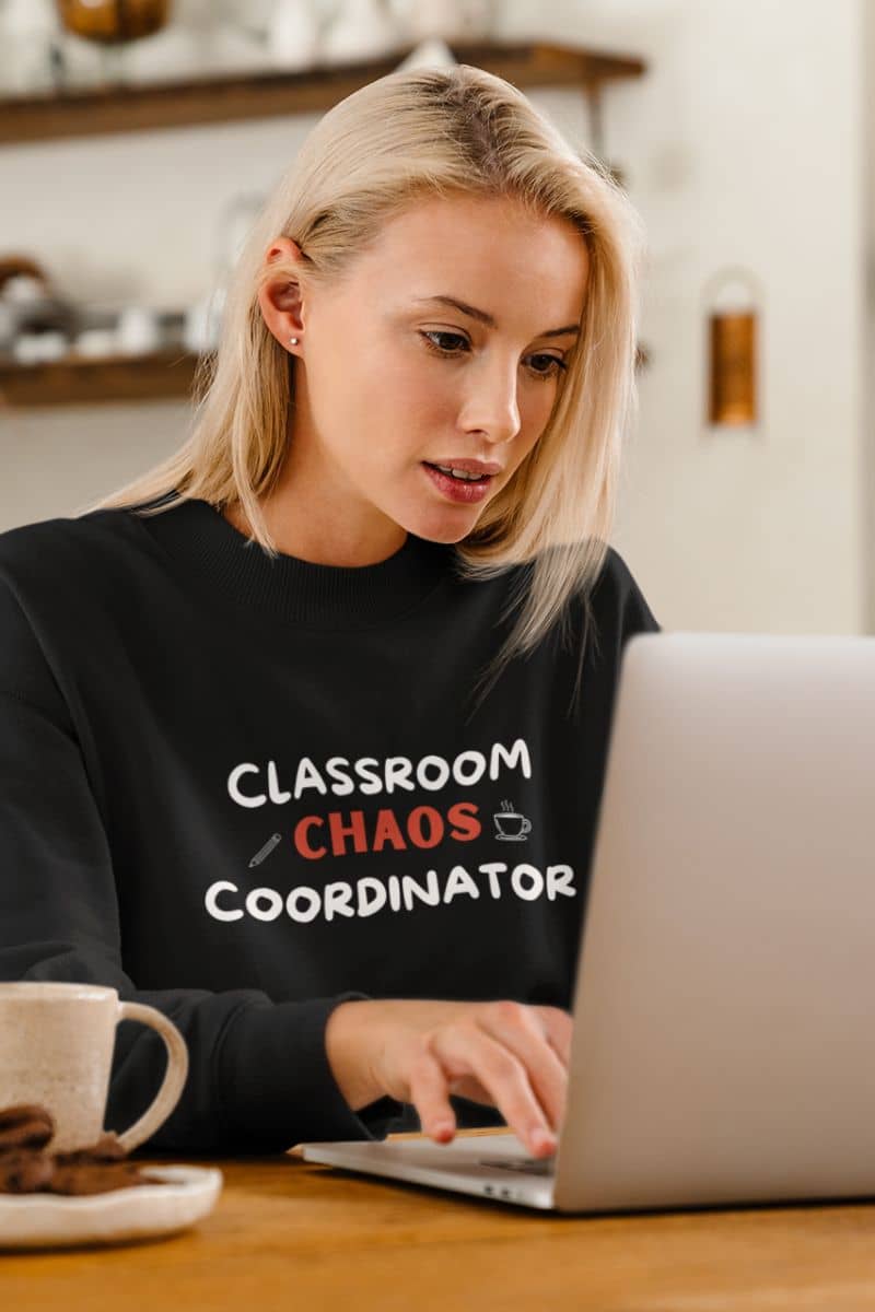 teacher appreciation sweatshirt with a blonde woman wearing a black crewneck sweatshirt with the text 'Classroom Chaos Coordinator' in white and red, typing on a laptop at a wooden table.