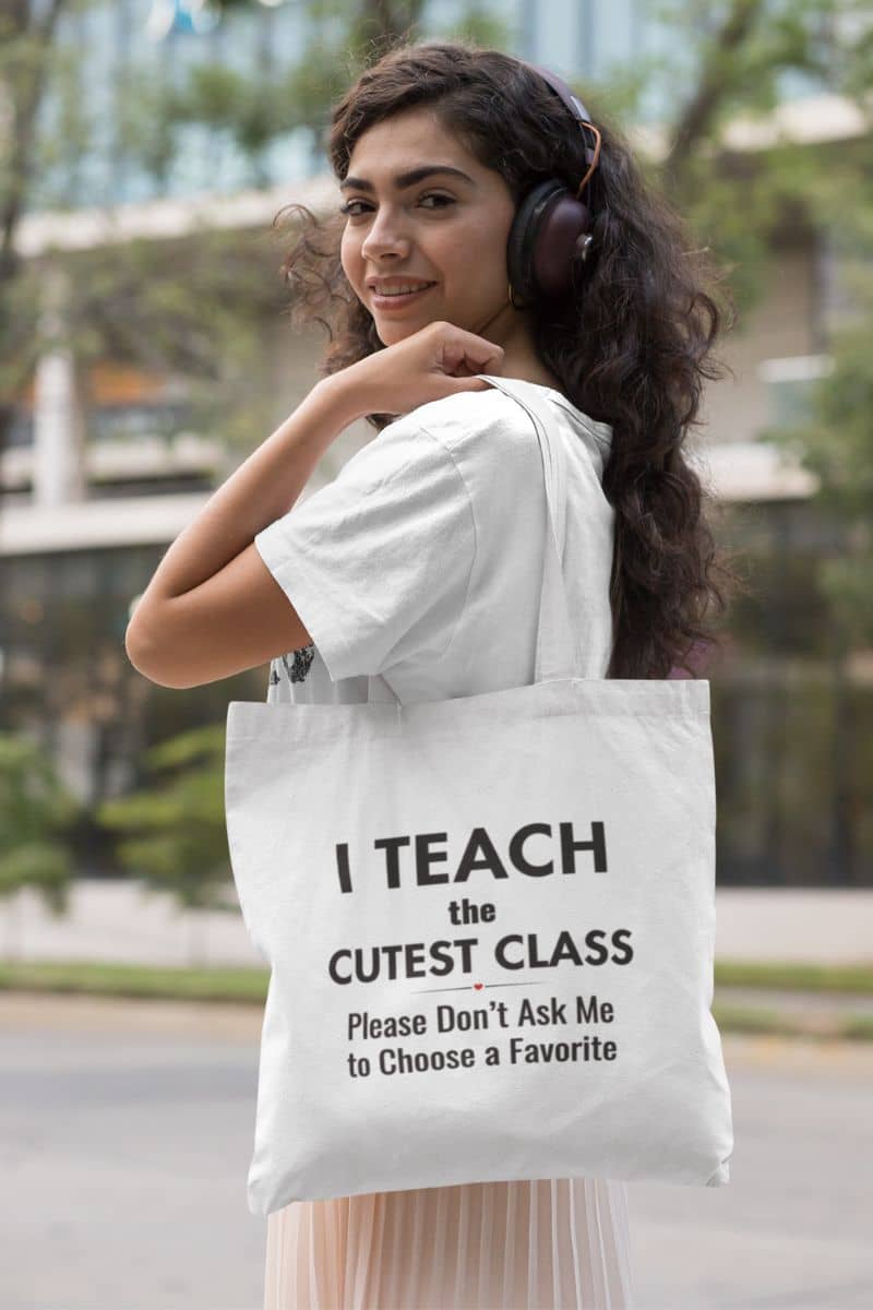 teacher canvas bags carried by a woman wearing headphones and a white t-shirt, featuring black text that reads 'I Teach the Cutest Class. Please Don’t Ask Me to Choose a Favorite' in an outdoor setting.