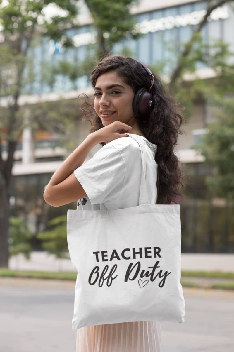 teacher carry bag with a woman wearing headphones and a white t-shirt carries a white canvas tote bag featuring the text 'TEACHER Off Duty' printed in black on the front, set against a blurred outdoor background.