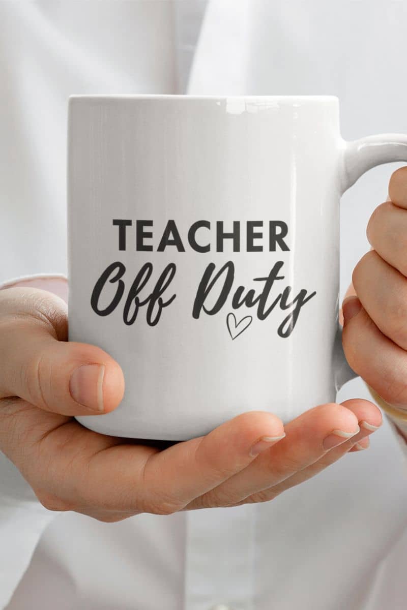 teacher coffee cup with a white ceramic mug featuring the text 'TEACHER Off Duty' in black lettering, held by two hands against a soft white background.