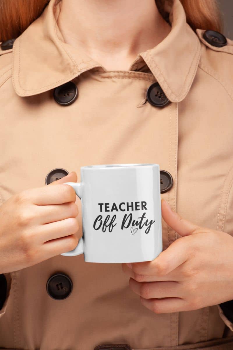 teacher coffee mug gift with a person in a beige trench coat holds a white ceramic mug featuring the text 'TEACHER Off Duty' printed in black on the front.
