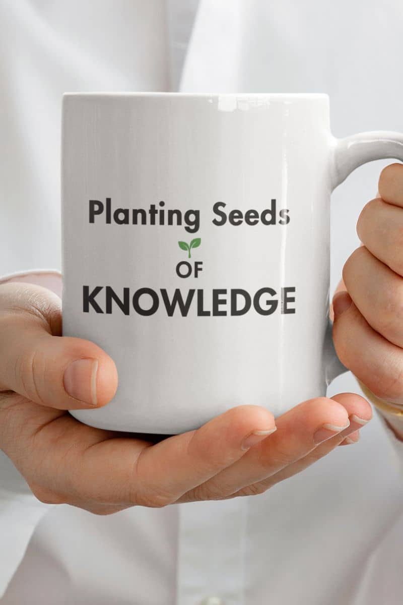 teacher coffee mug with a white mug held in two hands against a white shirt background. The mug features the printed text 'Planting Seeds OF KNOWLEDGE' 