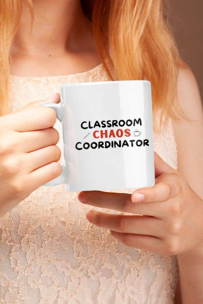teacher coffee mug with a person in a light beige lace top holds a white ceramic mug with the text 'Classroom Chaos Coordinator' printed on it. The text features black and red lettering with small graphic accents, set against a warm, softly blurred background.