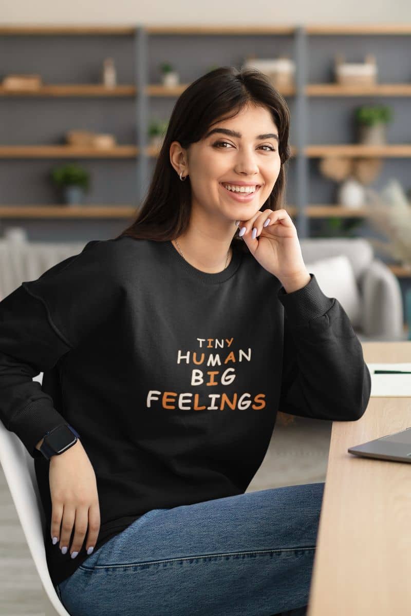 teacher crewneck sweatshirt with a woman wearing a black sweatshirt featuring the text 'TINY HUMAN BIG FEELINGS' 
