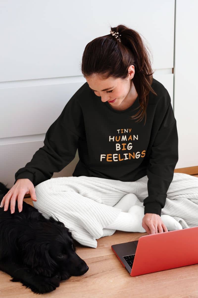 teacher crewneck with a woman wearing a black sweatshirt with text 'TINY HUMAN BIG FEELINGS' in gold and white lettering, sitting on a wooden floor with a black dog and a red laptop