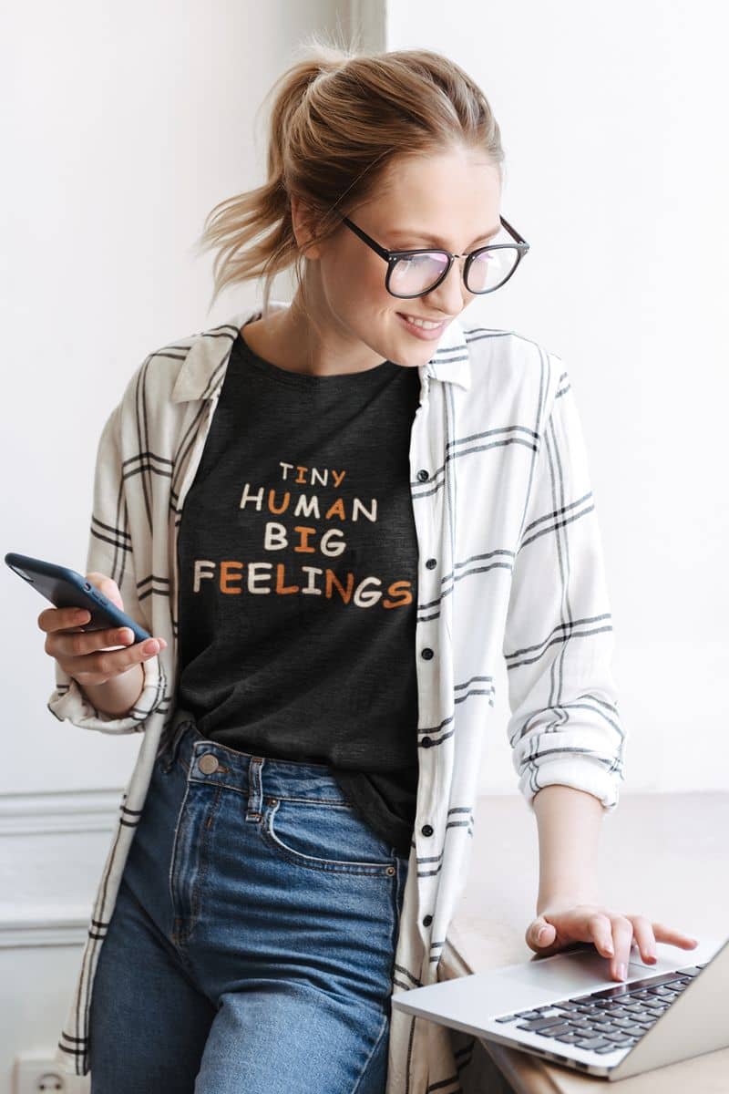 teacher graphic tee with a woman wearing a black graphic t-shirt with the text 'Tiny Human Big Feelings' in gold and orange, layered under an open white and gray plaid flannel shirt. She is also wearing blue jeans and glasses, holding a smartphone and using a laptop.