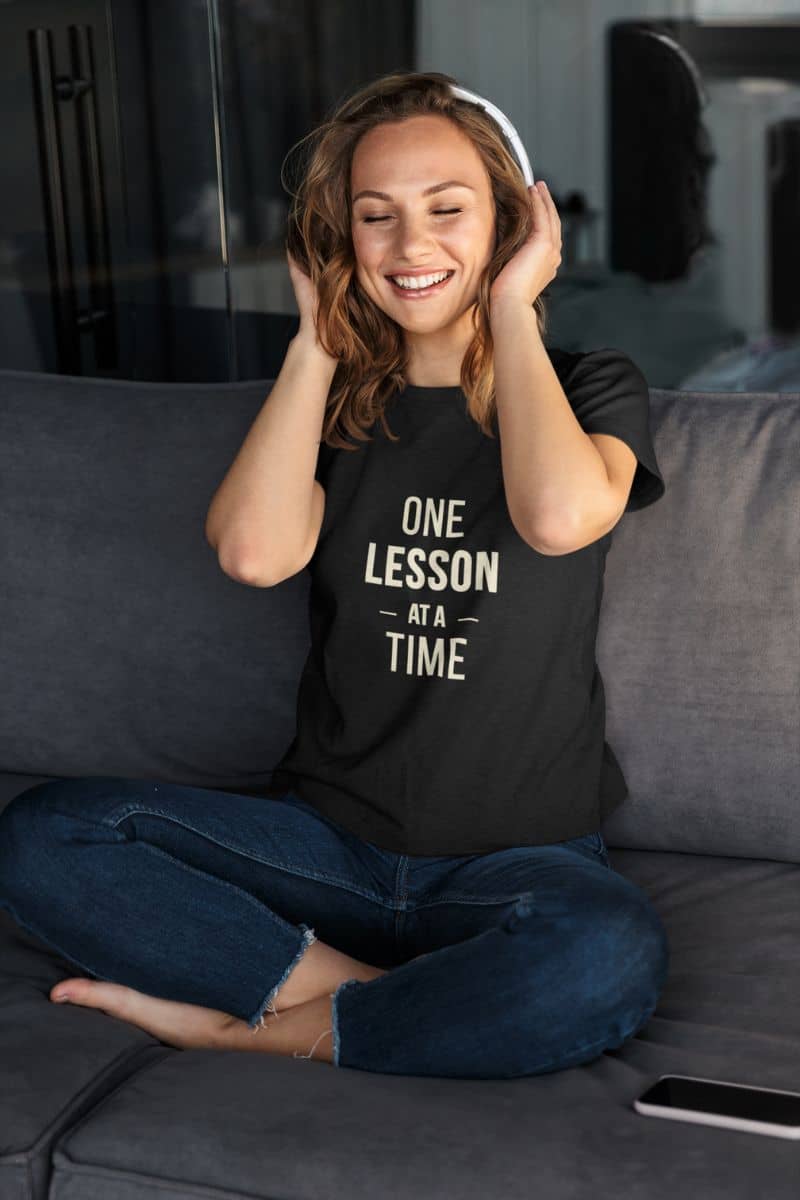teacher graphic tee with a woman sitting cross-legged on a gray couch, wearing a black t-shirt featuring the text 'ONE LESSON AT A TIME'.