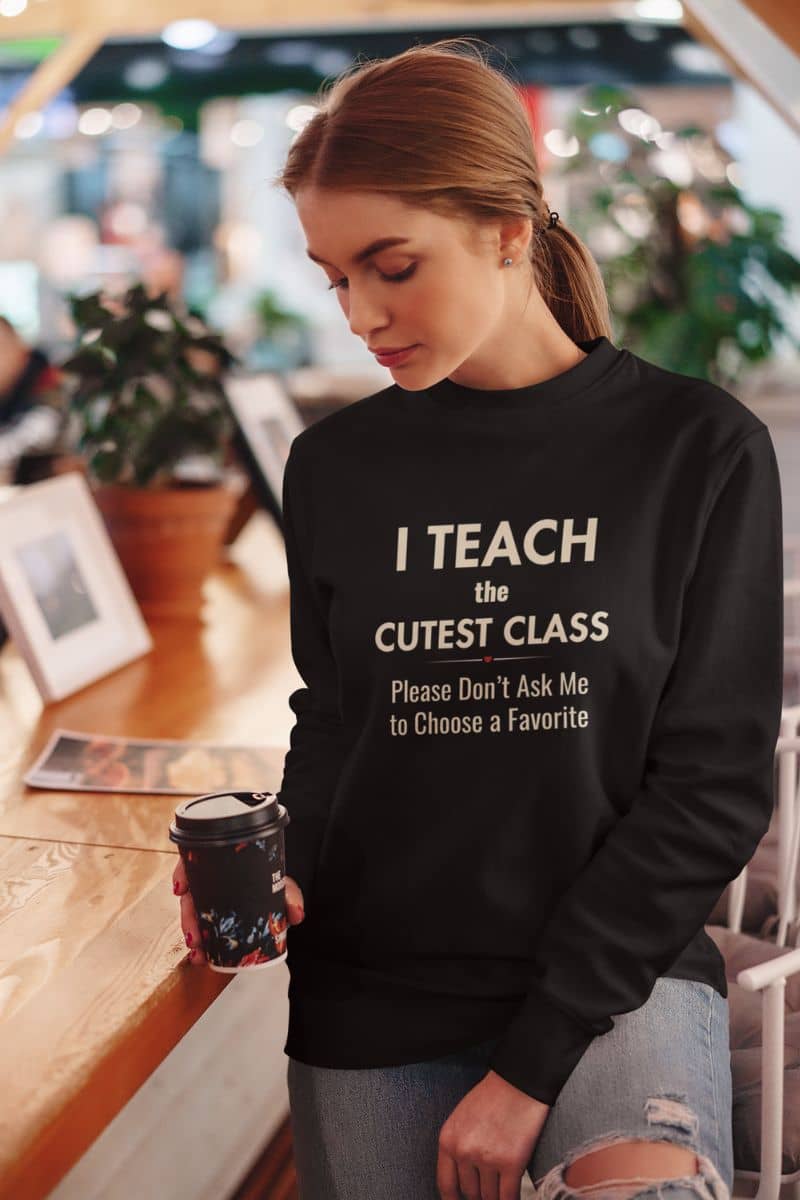 teacher pullover with a woman wearing a black crewneck sweatshirt featuring the text 'I Teach the Cutest Class. Please Don’t Ask Me to Choose a Favorite', standing in a cafe setting and holding a coffee cup.