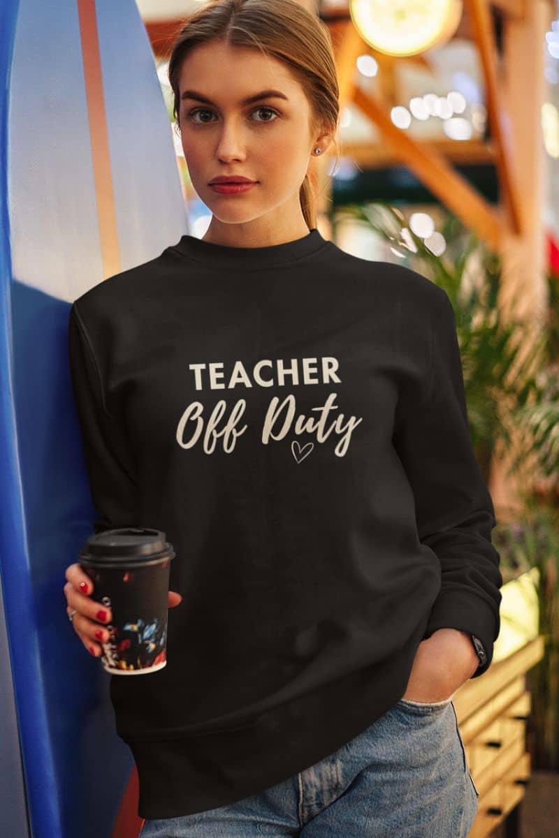 teacher pullover with a woman wearing a black crewneck sweatshirt featuring the text 'TEACHER Off Duty', holding a coffee cup and standing next to a blue surfboard in a warm, bokeh-filled indoor setting.