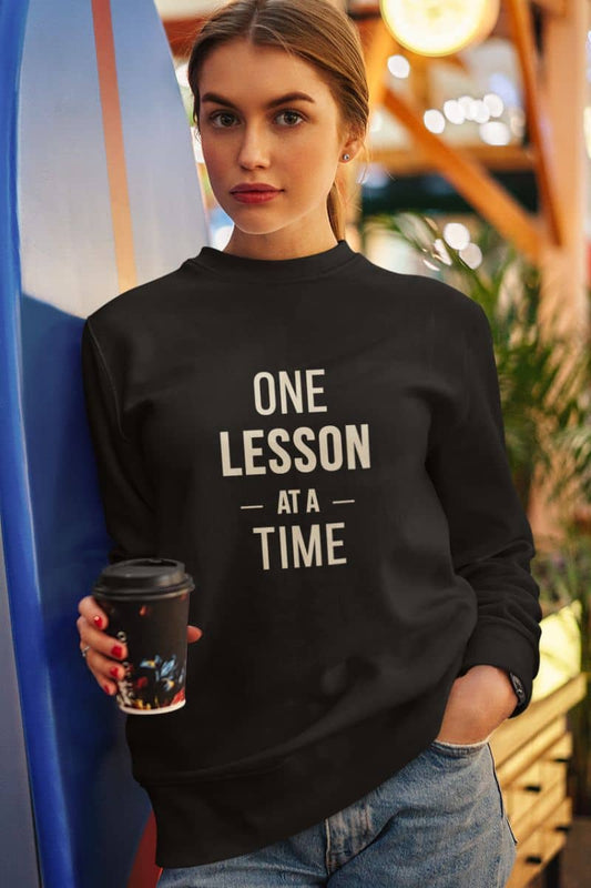 teacher pullover with a woman wearing a black sweatshirt featuring the beige printed text 'ONE LESSON AT A TIME' holds a coffee cup while leaning against a blue surfboard in a cafe setting.