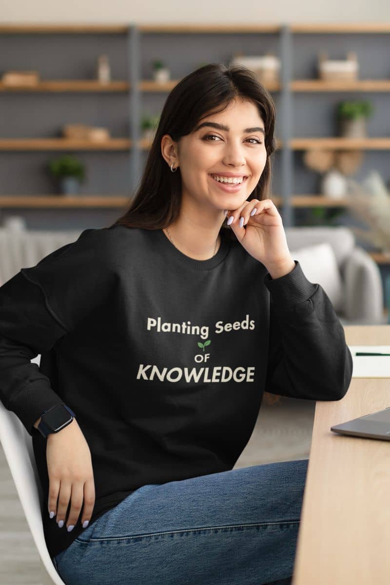 teacher pullover with a woman sitting in a chair at a desk, wearing a black sweatshirt featuring the text 'Planting Seeds of KNOWLEDGE'