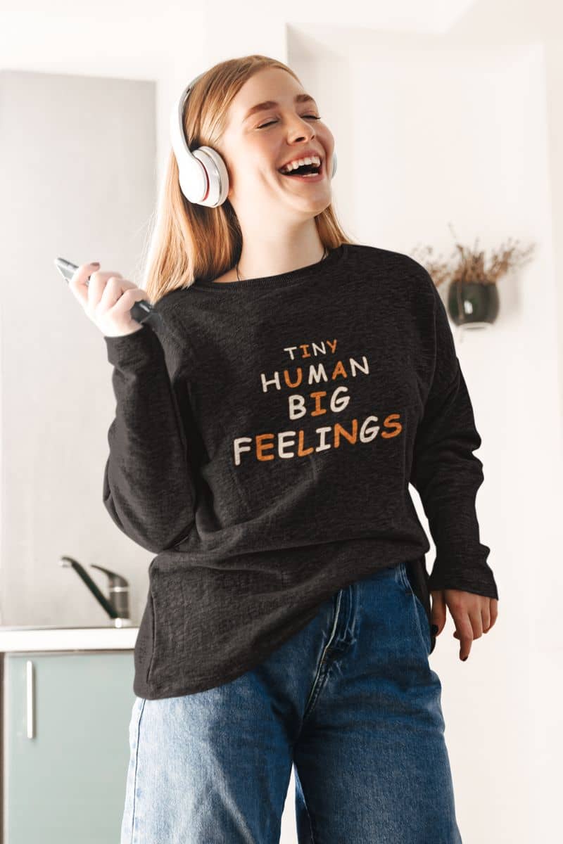 teacher sweatshirt with a woman wearing a dark charcoal heather long-sleeve sweatshirt with text 'Tiny Human Big Feelings' 