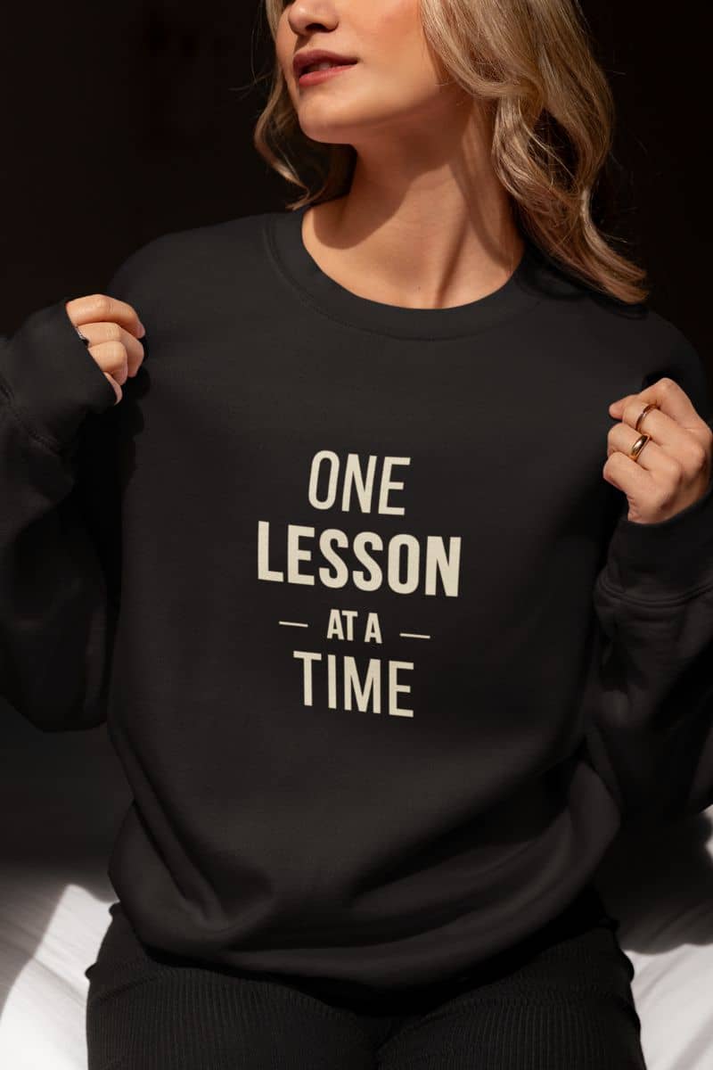 teacher sweatshirt with a woman wearing a black crewneck sweatshirt featuring the text 'ONE LESSON AT A TIME', holding the shoulders of the sweatshirt.