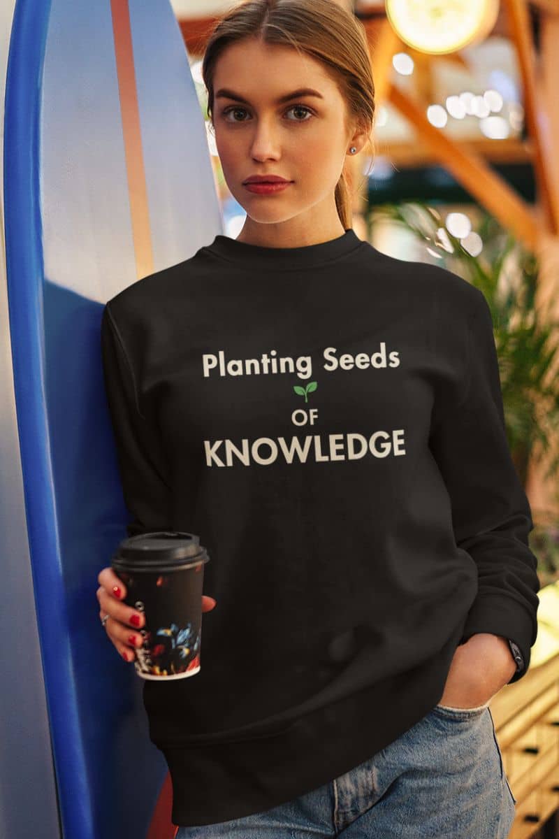 teacher sweatshirt with a woman wearing a black sweatshirt featuring the text 'Planting Seeds OF KNOWLEDGE' and holding a coffee cup, standing next to a blue surfboard in a warm, bokeh-filled indoor setting.