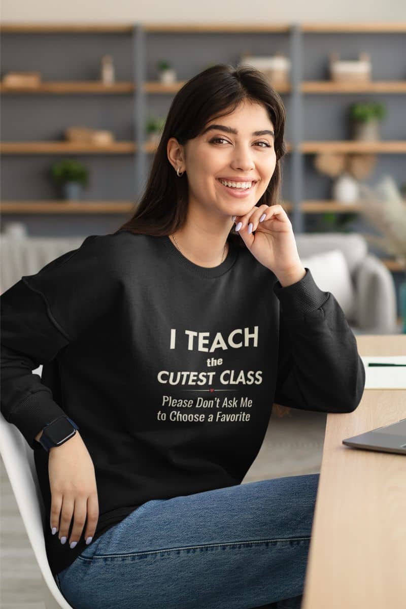 teacher sweatshirt with a woman wearing a black crewneck sweatshirt featuring the text that reads 'I Teach the Cutest Class. Please Don’t Ask Me to Choose a Favorite', paired with blue jeans, sitting in a modern office setting.