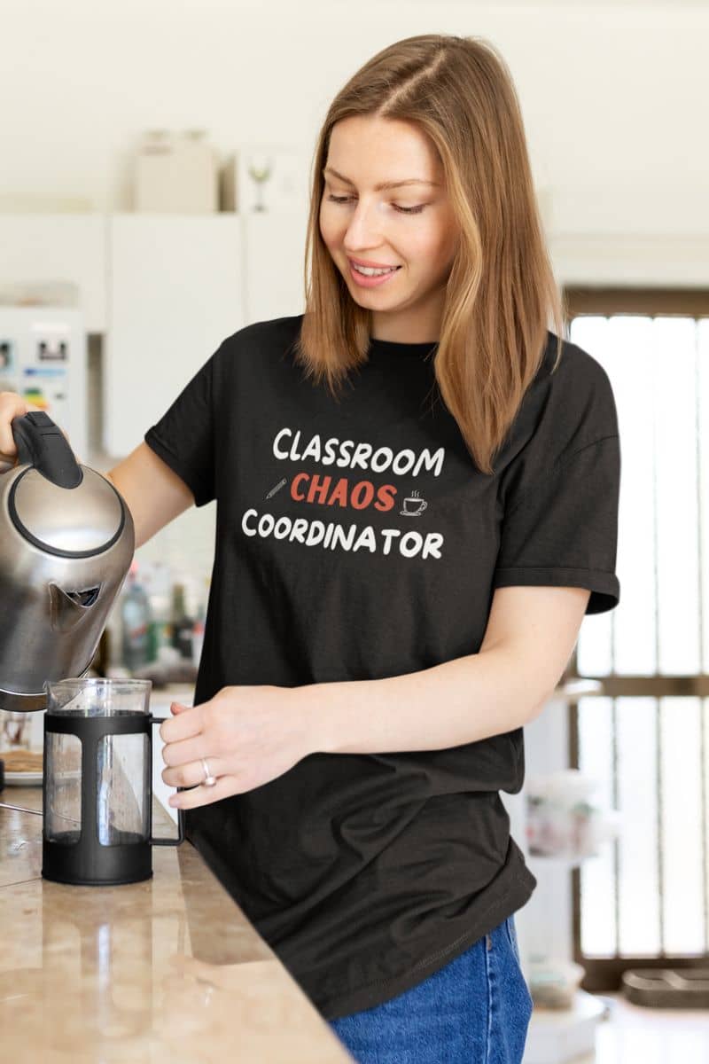 teacher t shirt with a woman in a kitchen wearing a black graphic t-shirt that reads 'Classroom Chaos Coordinator' in white and orange text. She is pouring water from a kettle into a French press.