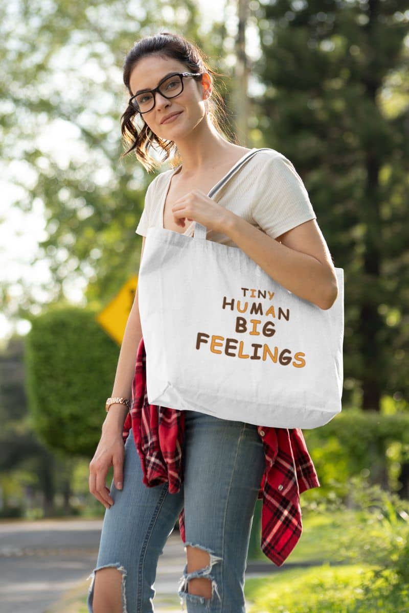 teacher tote bag with a woman carrying a white canvas tote bag with text 'TINY HUMAN BIG FEELINGS', outdoors