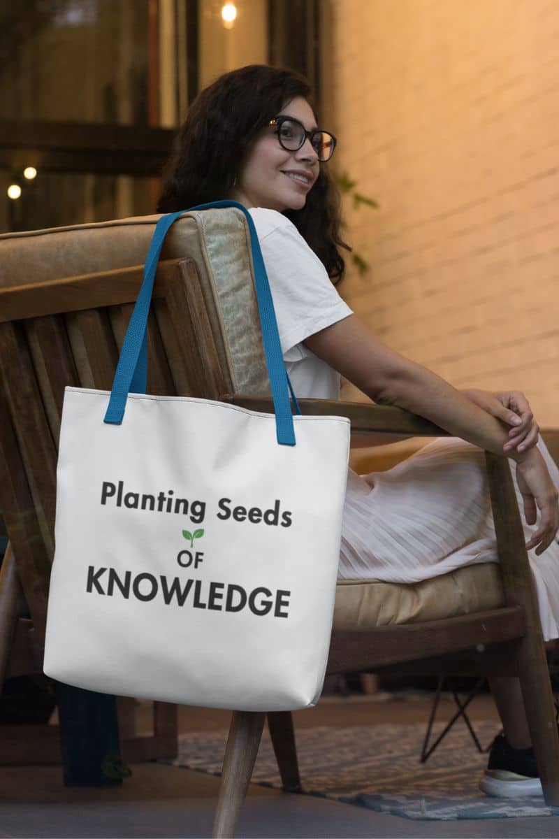 teacher tote bag with a white canvas tote bag featuring the text 'Planting Seeds OF KNOWLEDGE', hanging on a wooden chair beside a woman sitting in a warm indoor setting 