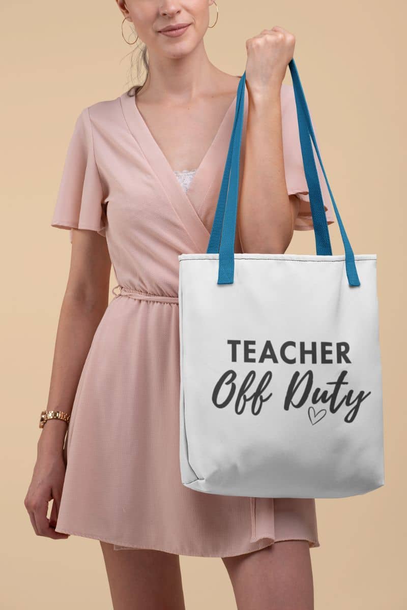 teacher tote for work with a woman in a light pink dress holds a white canvas tote bag with blue shoulder straps. The bag features the text 'TEACHER Off Duty' in black script and print, with a small heart below.