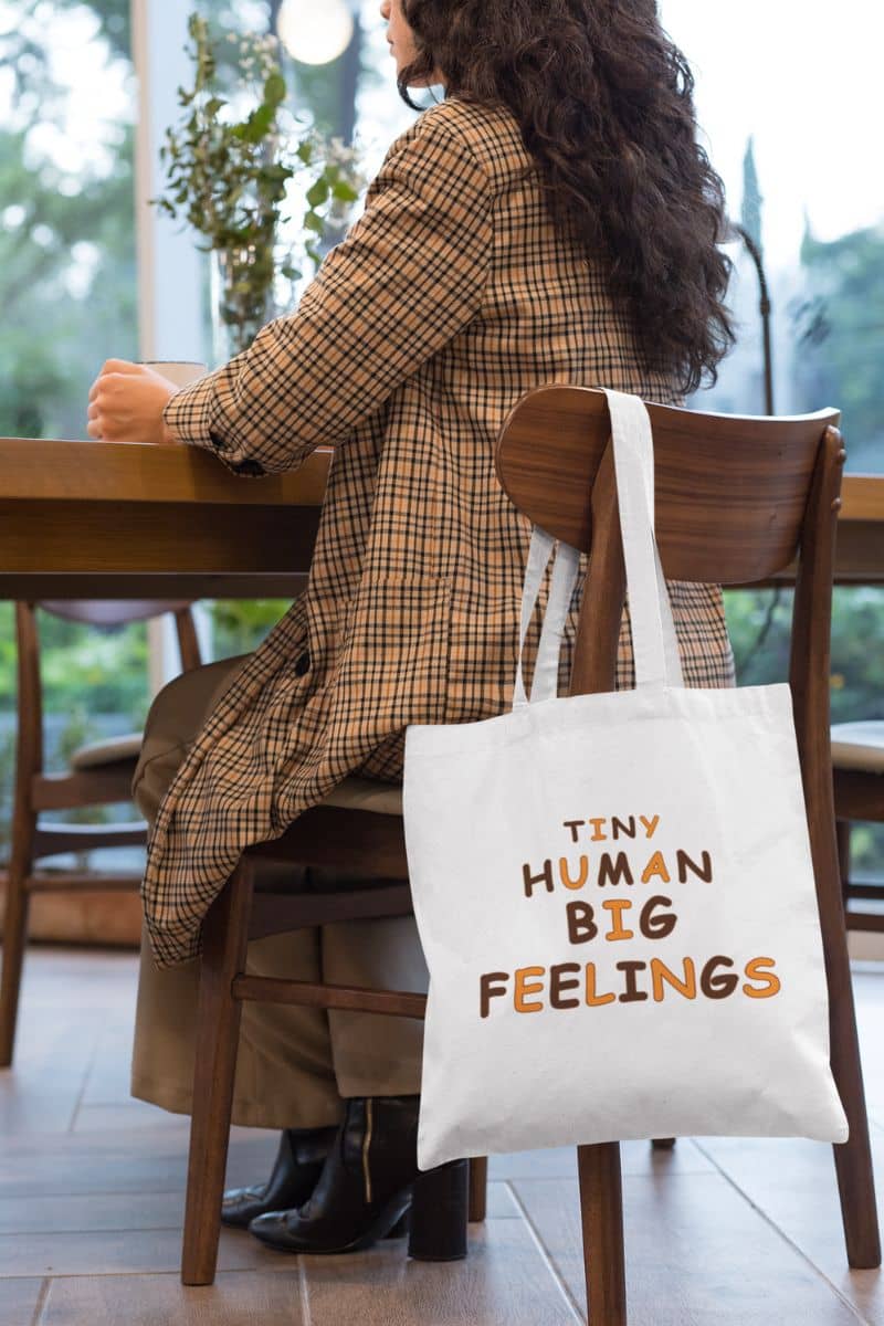 teacher tote featuring a white canvas tote bag with text 'TINY HUMAN BIG FEELINGS', hanging from a wooden chair next to a woman in a brown plaid coat seated at a wooden table in a cafe.