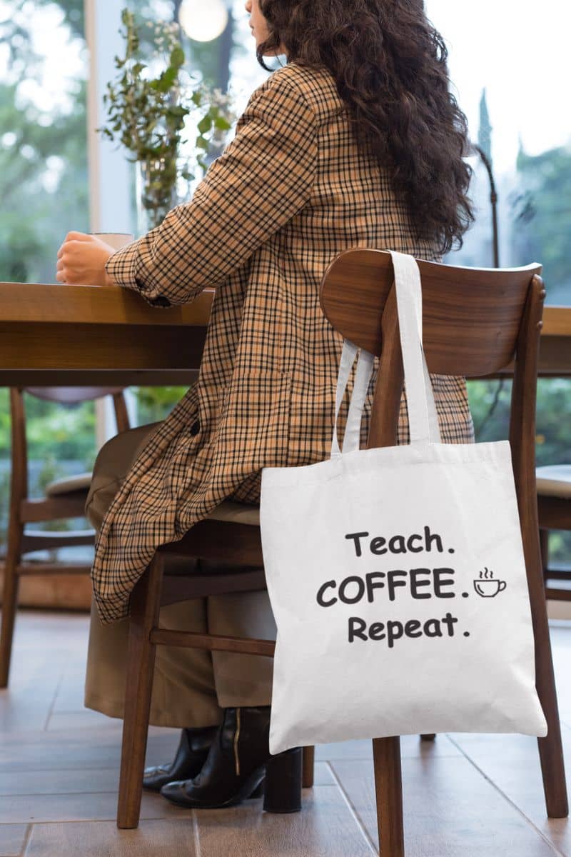 teacher work bag with a white canvas tote bag featuring the text 'Teach. Coffee. Repeat.', hanging on the chair in the coffee shop