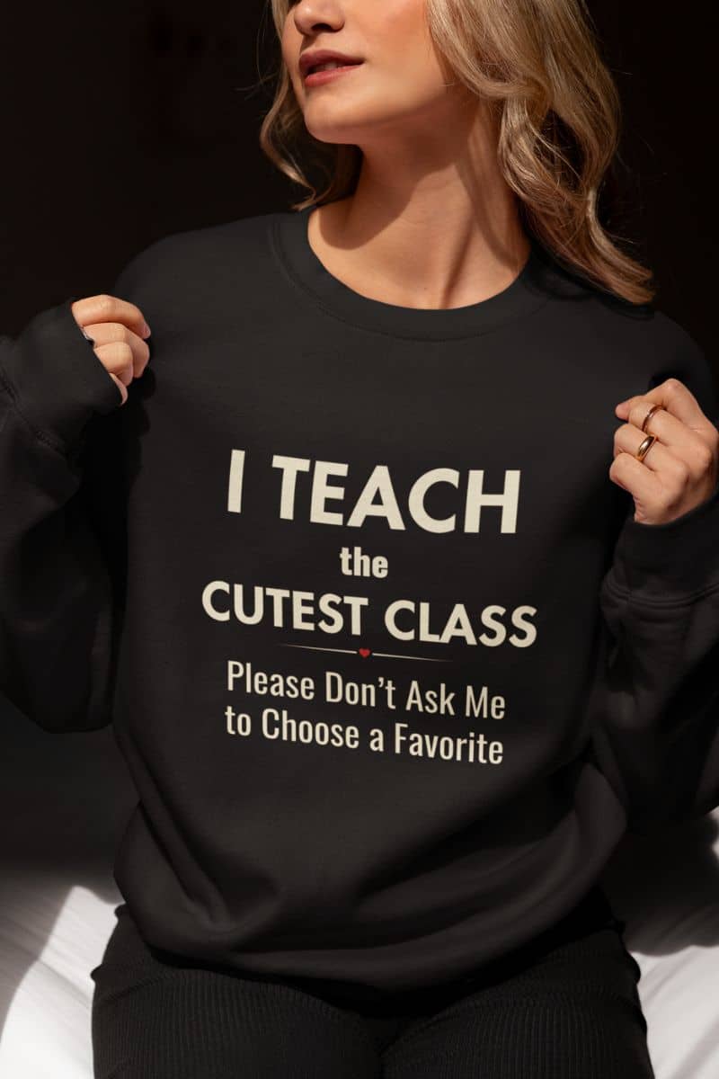 teachers sweater with a woman wearing a black crewneck sweatshirt featuring the text 'I Teach the Cutest Class. Please Don’t Ask Me to Choose a Favorite' printed in white, against a dark background.