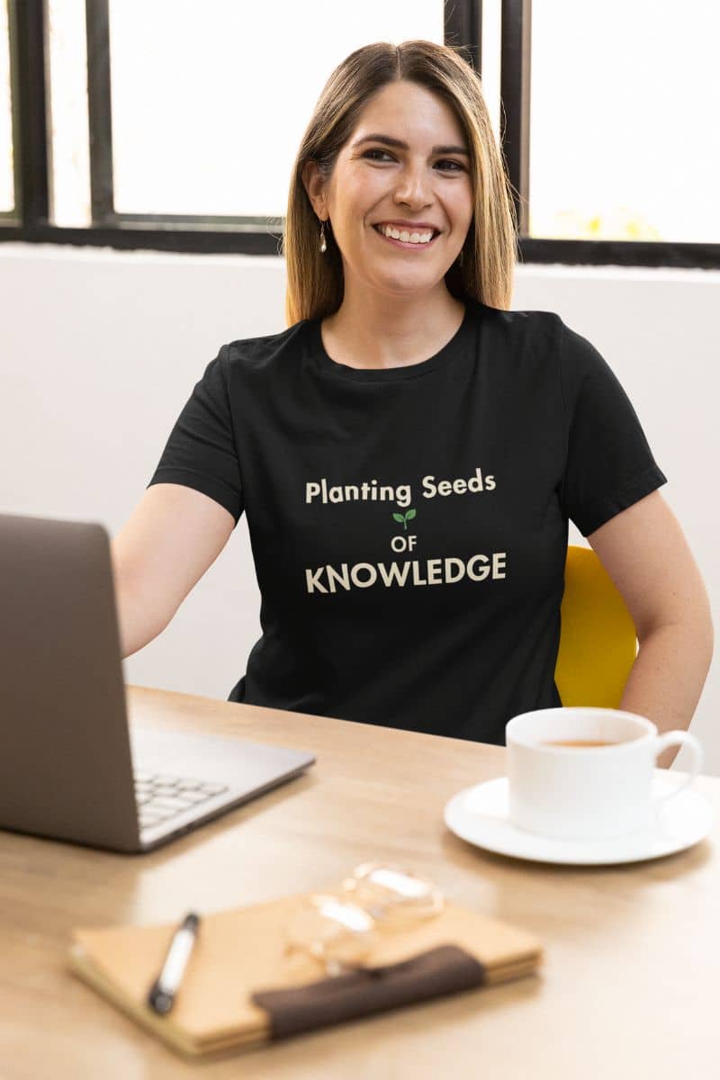 teaching shirt with a woman wearing a black t-shirt that reads 'Planting Seeds OF KNOWLEDGE', sitting at a wooden desk