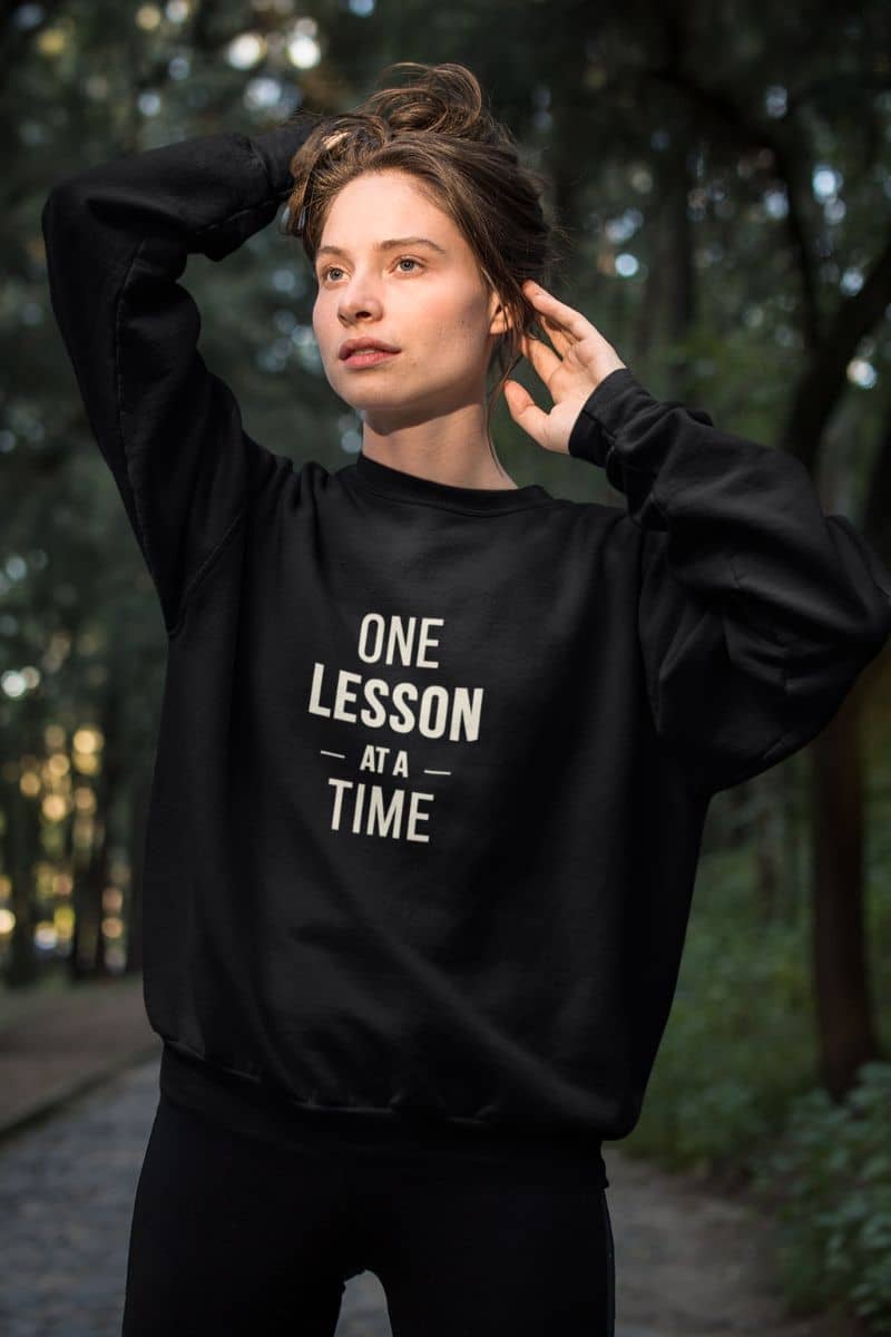 teaching sweatshirt with a woman wearing a black crewneck sweatshirt featuring the text 'ONE LESSON AT A TIME' printed in white on the front, standing outdoors on a path surrounded by trees.