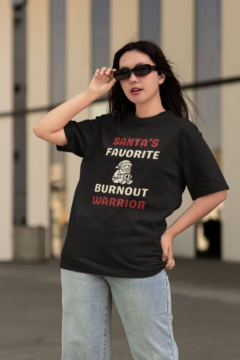 tired adult christmas tee with a person wearing a black t-shirt featuring the text  'Santa's Favorite Burnout Warrior' and graphic, standing outdoors.