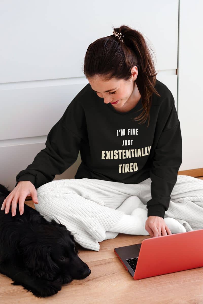 tired but trying sweatshirt with a woman sitting on the floor with a laptop and a dog, wearing a black sweatshirt with text 'I’m fine. Just existentially tired.'.
