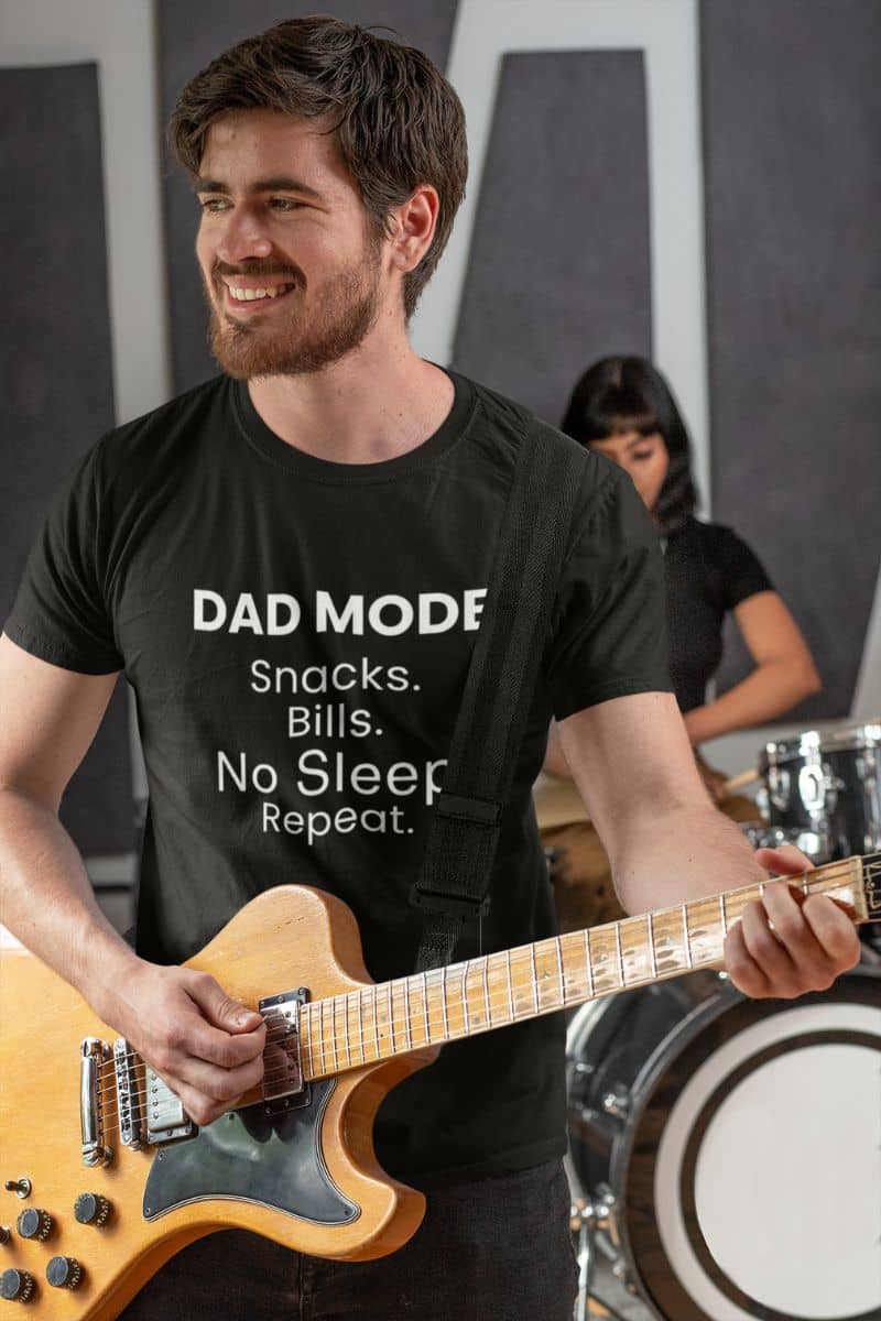tired dad shirt with a man playing guitar wearing a black t-shirt with text 'DAD MODE:  Snacks. Bills. No Sleep. Repeat.', with a blurred background.