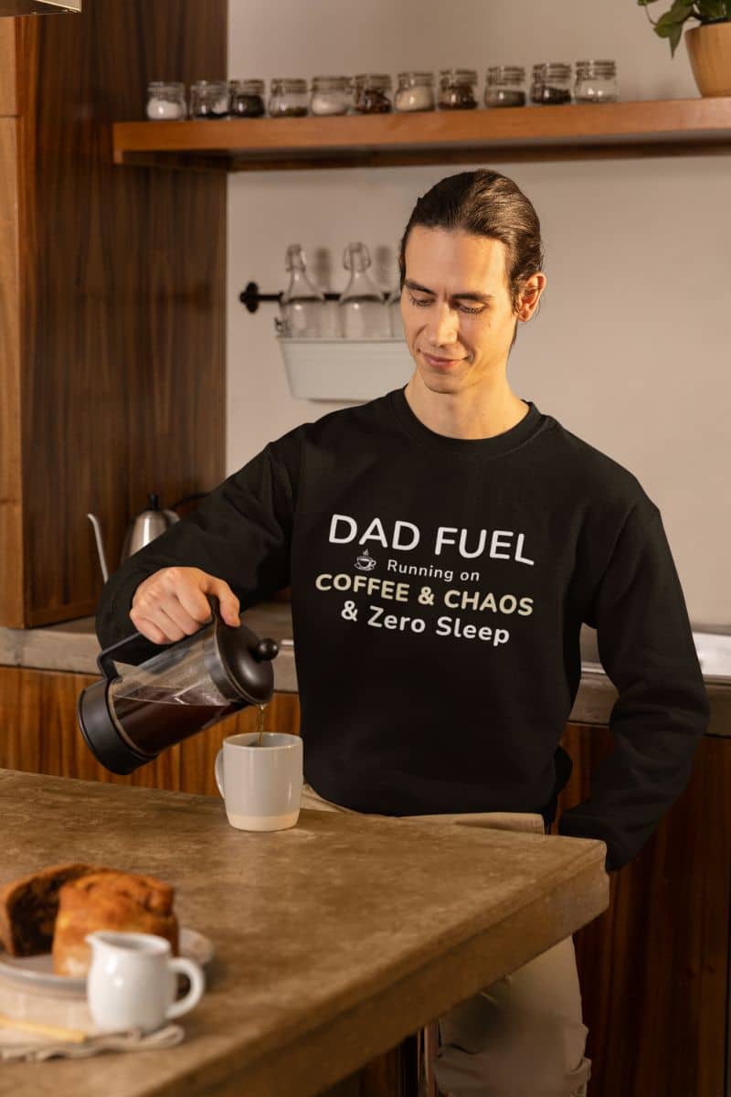 tired dad sweatshirt with a person pouring coffee wearing a black sweatshirt with text 'DAD FUEL – Running on Coffee & Chaos & Zero Sleep', standing in a kitchen.