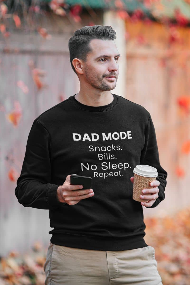 tired dad sweatshirt with a man wearing a black sweatshirt with text 'DAD MODE:  Snacks. Bills. No Sleep. Repeat.', holding a coffee cup and phone, with a blurred autumn background.
